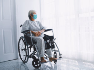 A sad elderly woman in a wheelchair wearing a surgical mask looking out of the window. In many cases, Iowa laws currently prevent residents of long term nursing facilities from seeing visitors, which raises the risk of nursing home neglect.